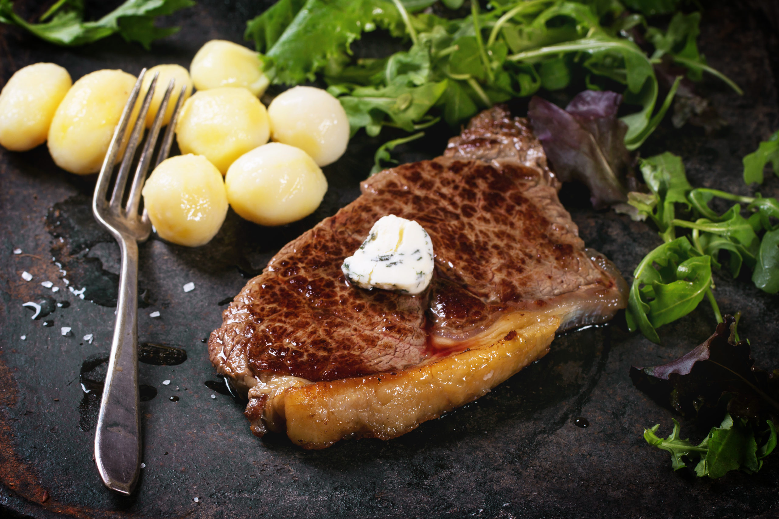 Grilled steak with butter, potatoes and green salad over black metal board